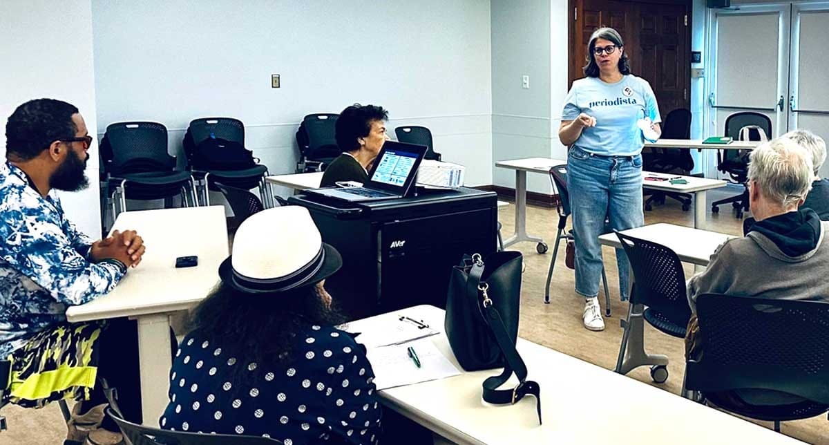 Photo showing a women wearing a shirt that says "periodista" and a button that says "The 51st" talking to a four people sitting at desks.