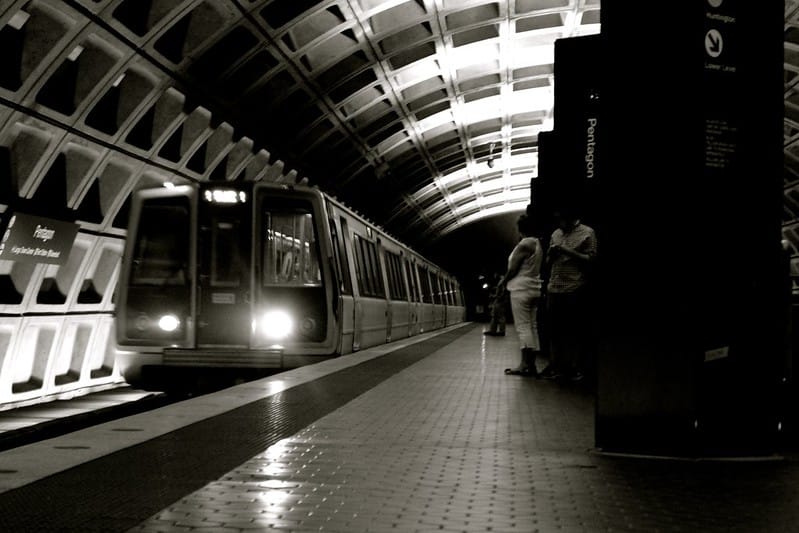 A metro train arrives to the Pentagon station while passengers wait on the platform. Photo in black and white.