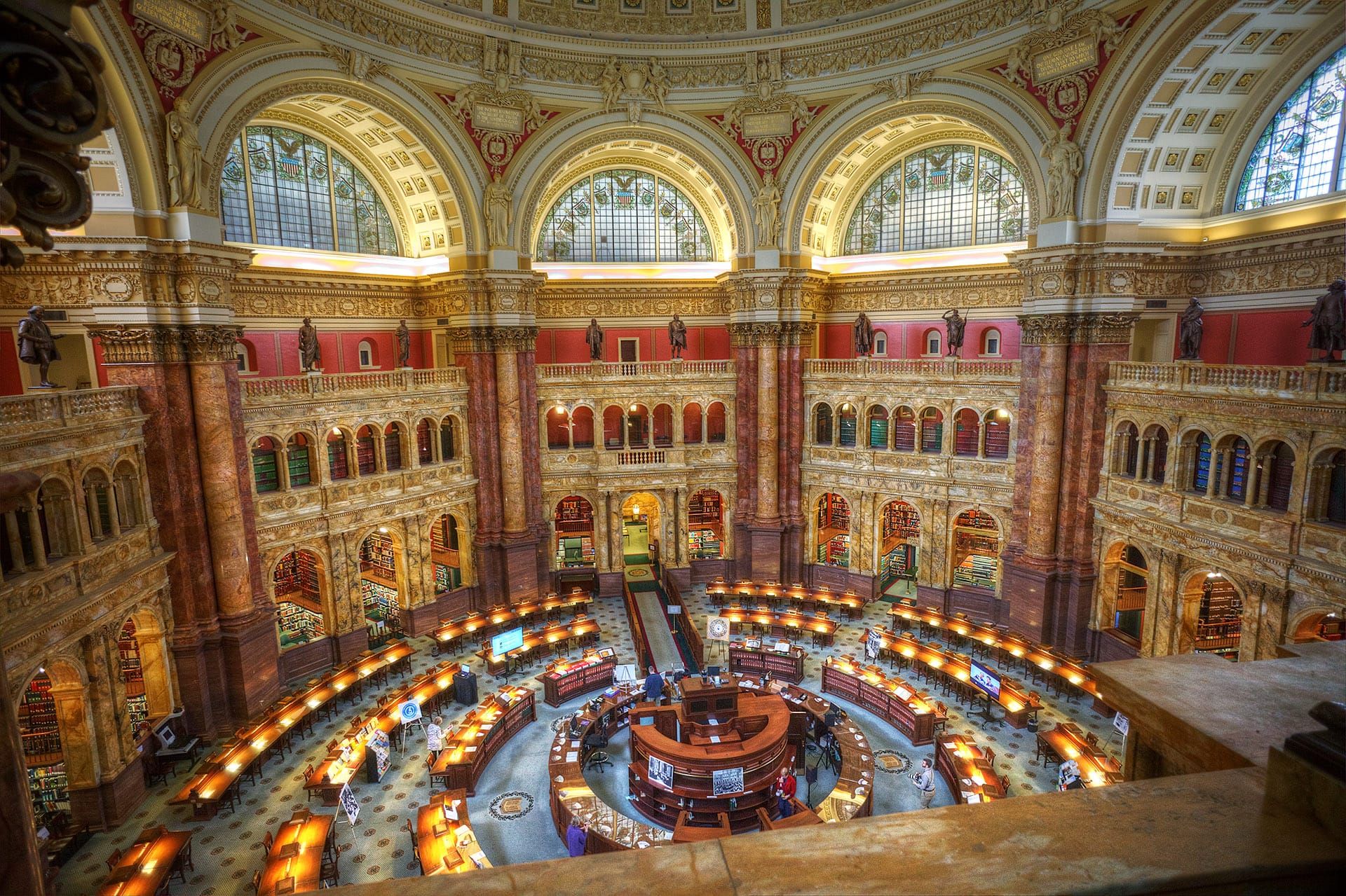 A photograph of the Library of Congress's stately Reading Room, which is ringed with desks and has large stained glass windows.