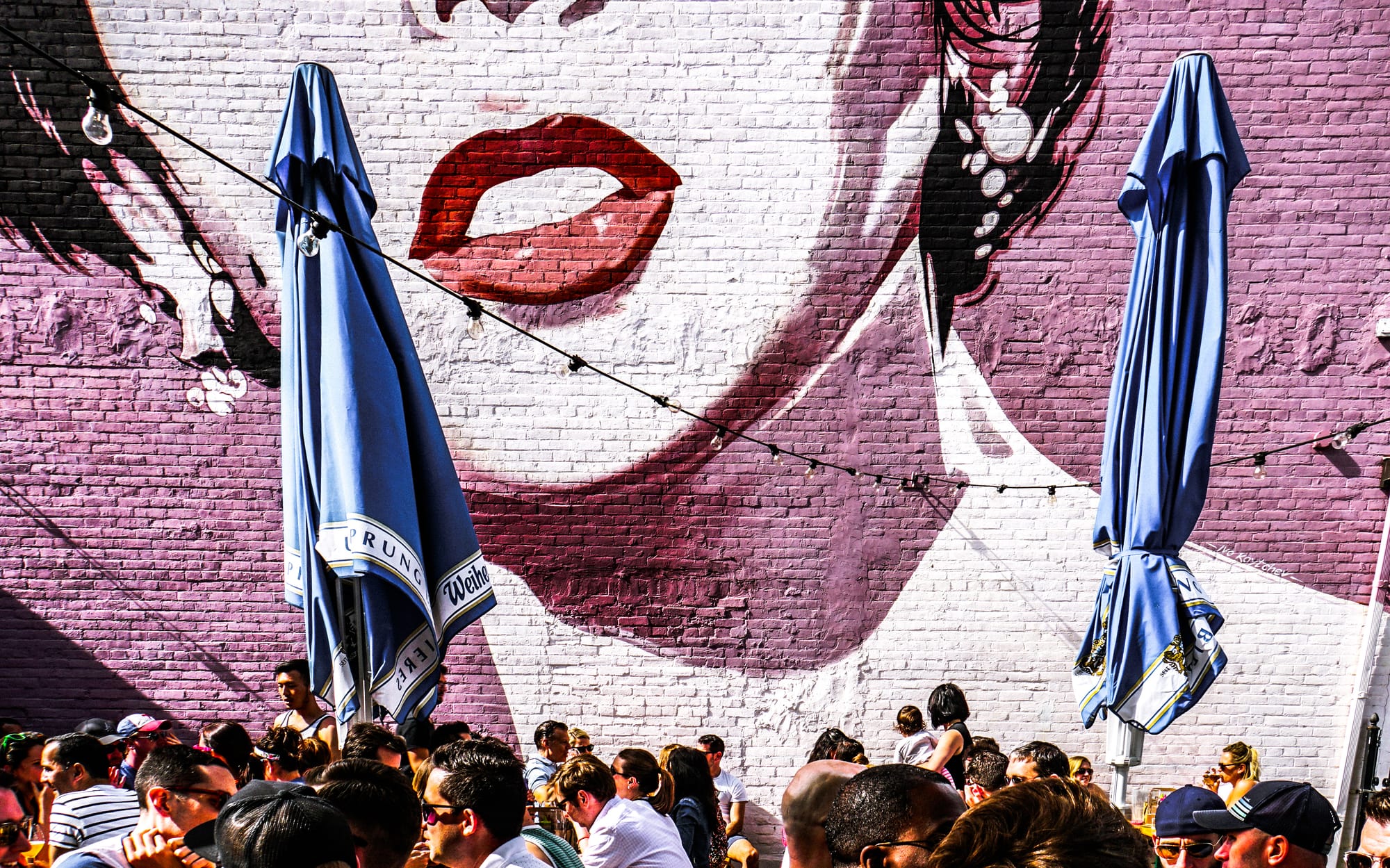 People gather around tables in an outdoor beer garden with two folded umbrellas and a large mural of a woman's face in the background.