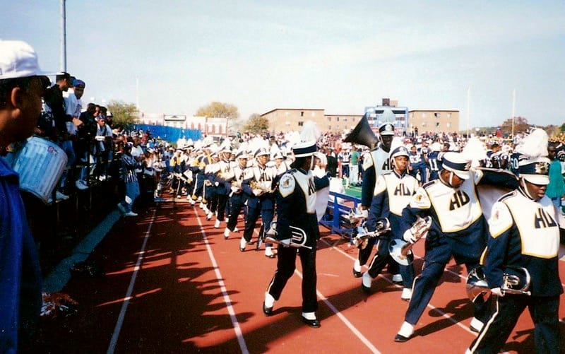 An older photograph of Howard University's marching band walking on the track next to a football field.