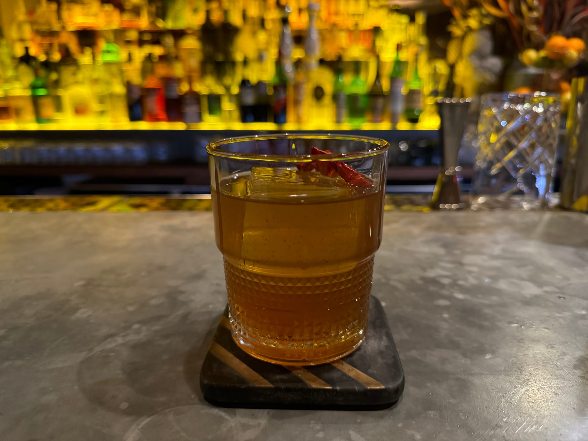 A brown cocktail in a glass sitting on a gray bar with liquor bottles in the background.