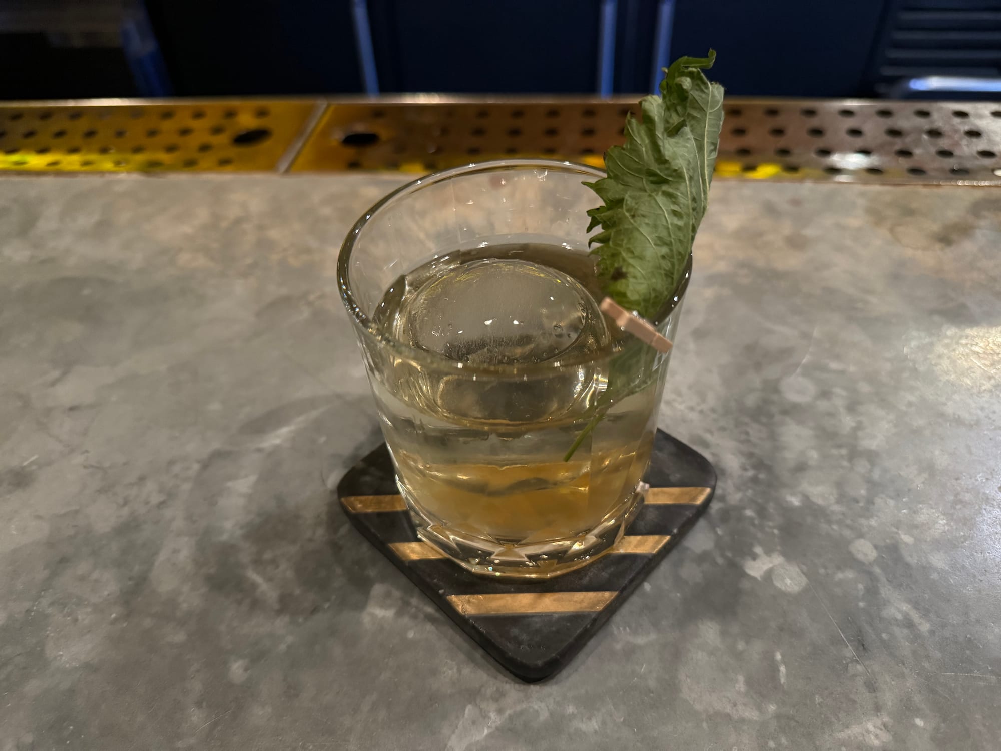 A circular glass sits on a bar tap. Inside, is a brown-ish drink with a large ice cube and green leaf.