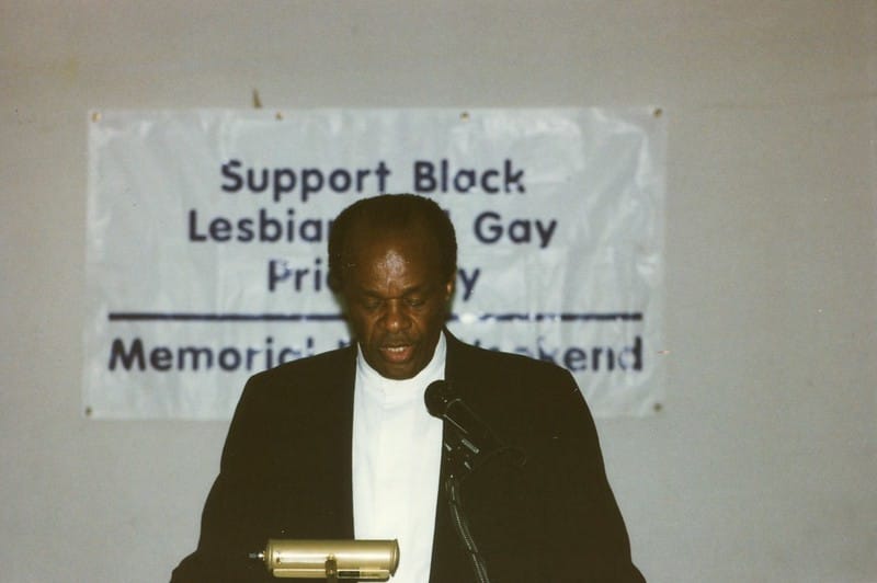 Marion Barry, an African American man, is speaking at a podium with a sign behind him supporting Black LGBTQ people.