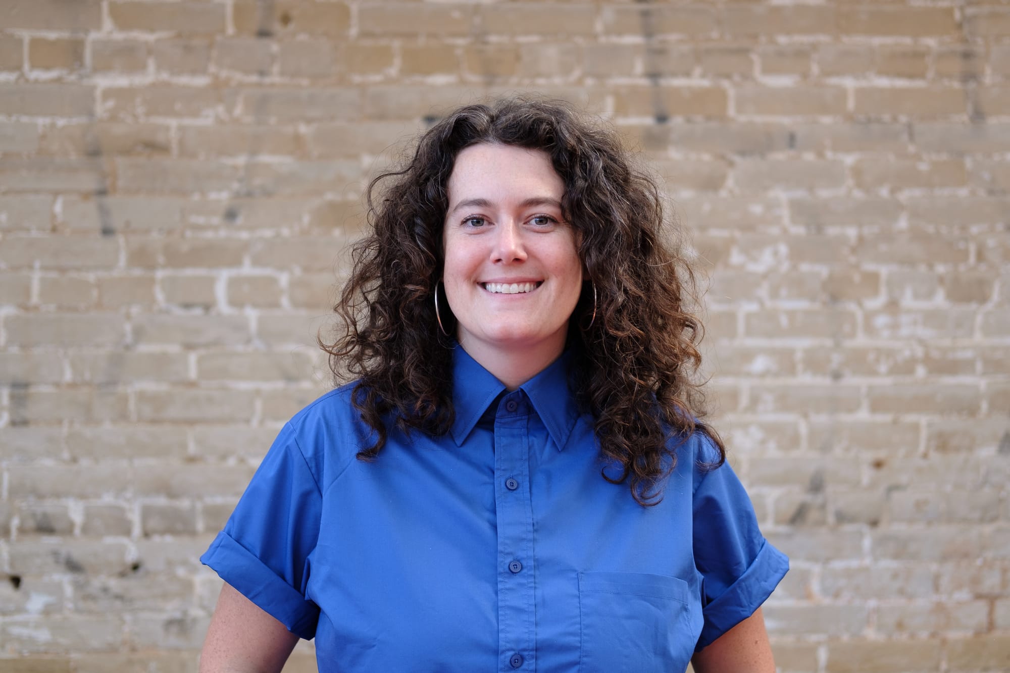 A photo of Maddie Poore, wearing a blue shirt with a beige brick background.