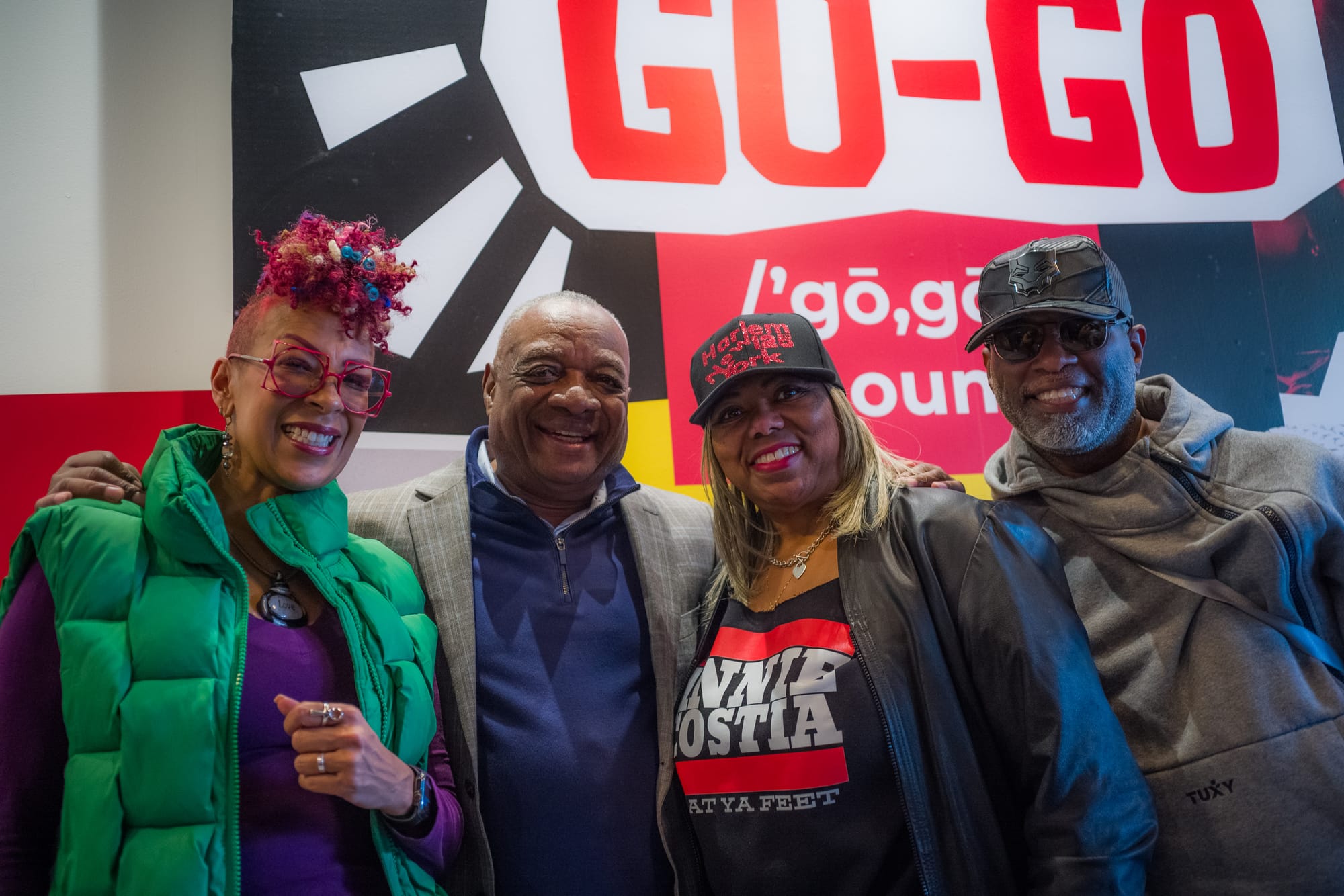 Two Black women and two Black men stand in front of a mural that reads "go-go."