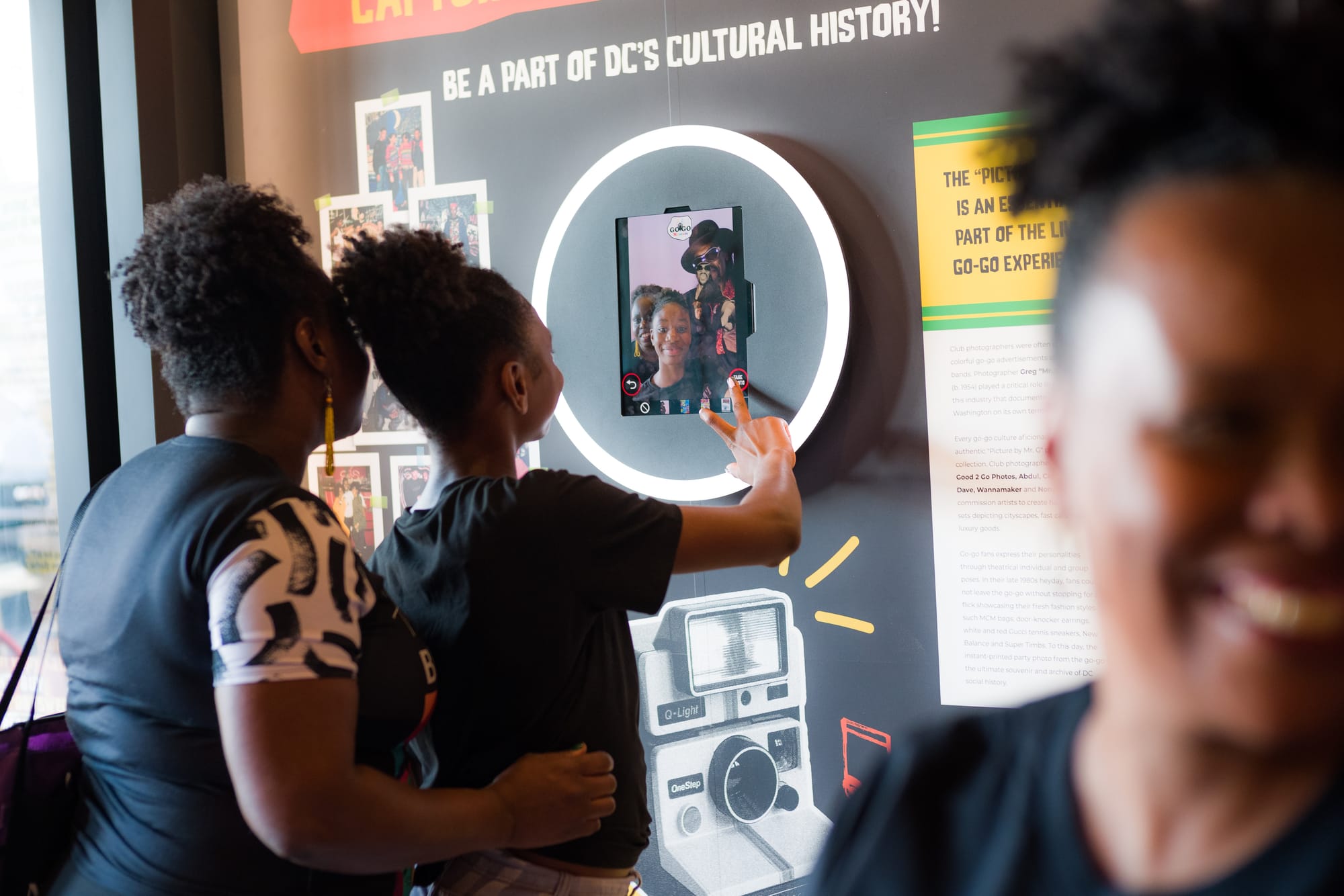 Two Black women stand in front of a selfie camera in an exhibit. 