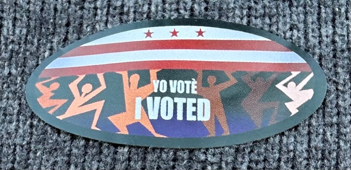 "I voted" sticker with an image of six figured in different skin tones holding up the D.C. flag (or perhaps being crushed by it)