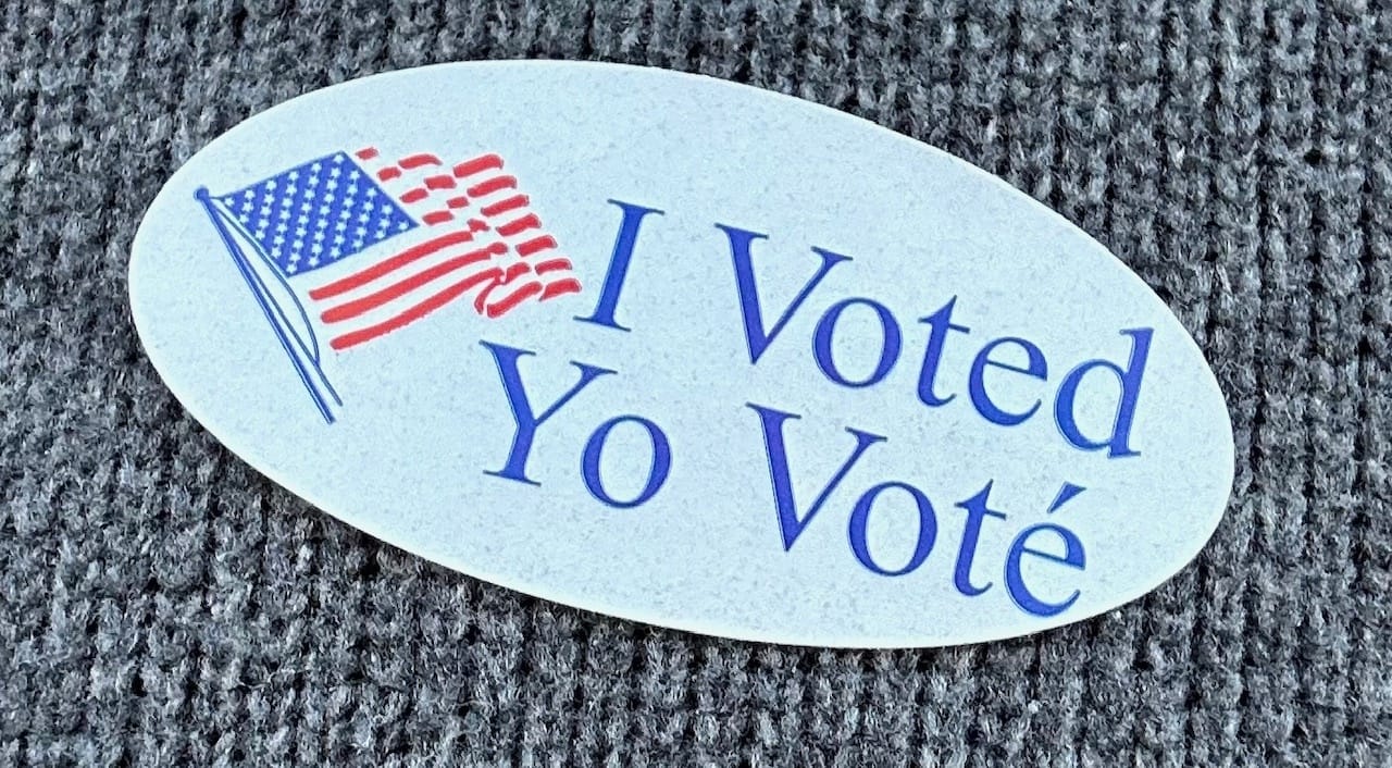 Classic "I voted" sticker with a white background, blue text, an American flag, and "I voted" written in both Spanish and English 