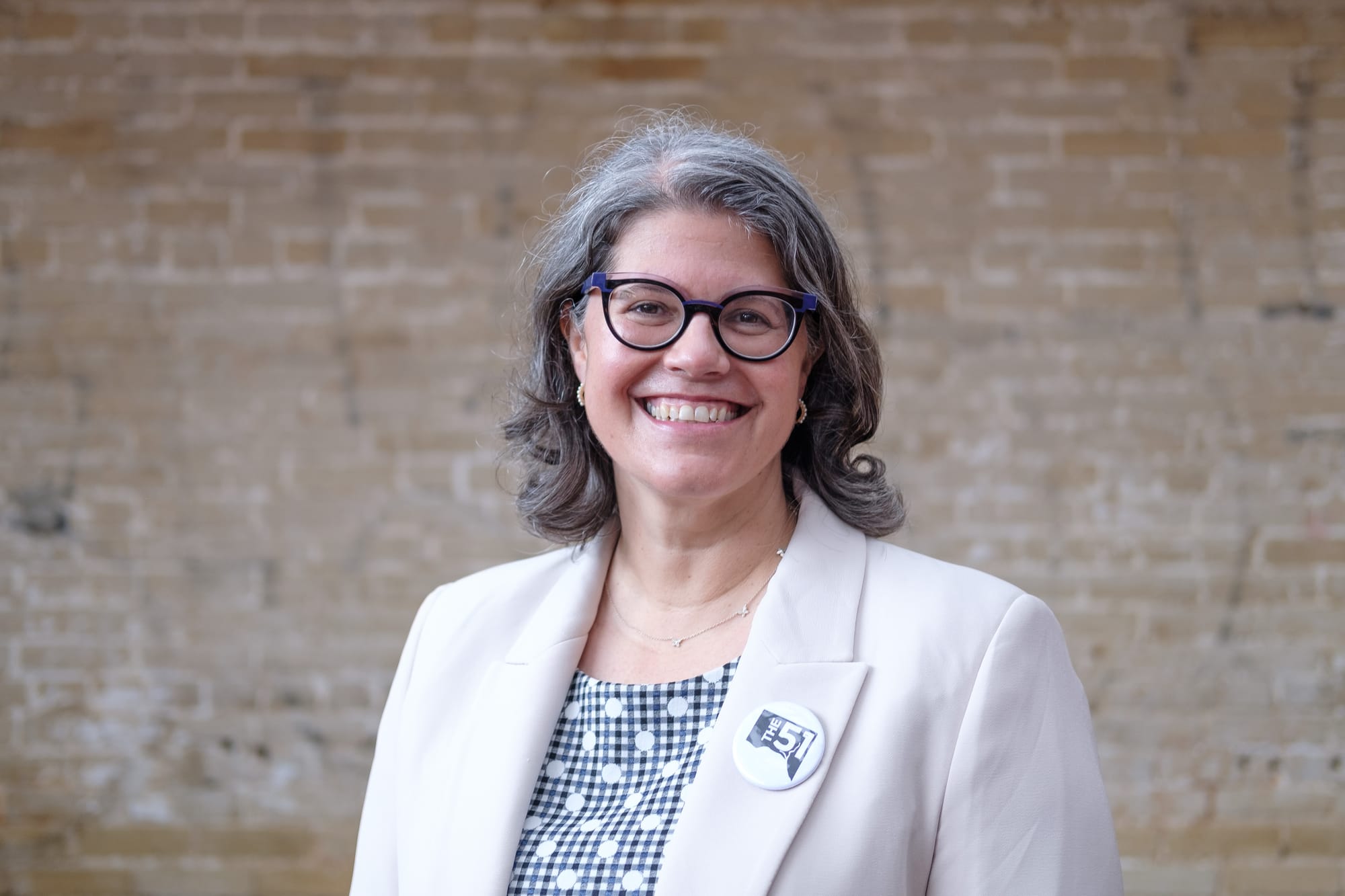 Woman in cream colored blazer, patterned blouse, and dark glasses stands, smiling, in front of a light-brown brick wall. 