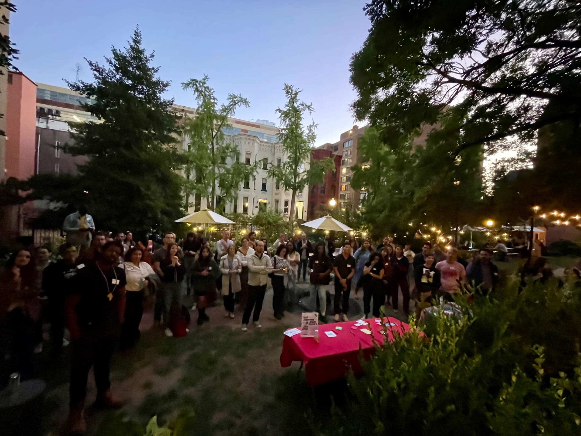 Photo showing many people gathered in a lit garden at dusk, looking toward the camera. A table with information about The 51st is visible in front of them.