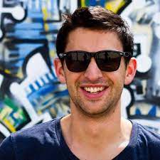 A photo of Tim, wearing sunglasses and standing in front of outdoor art.