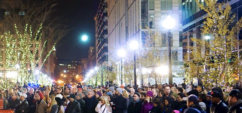 Large crowd gathers outside in downtown DC with holiday lights in the background.