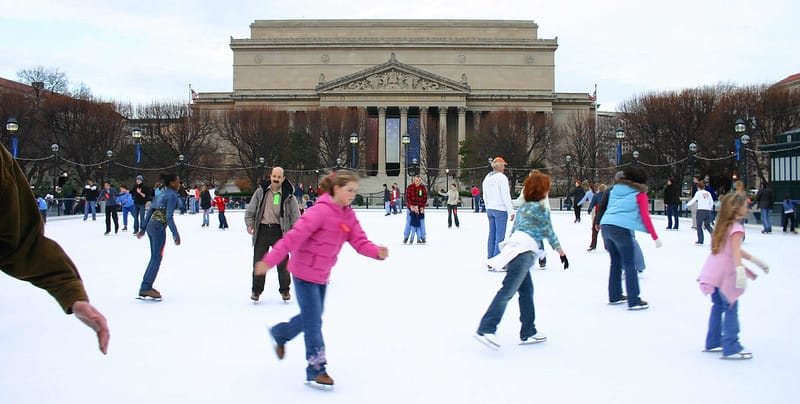 More than a dozen children and adults ice skating outdoors with a museum in the background.