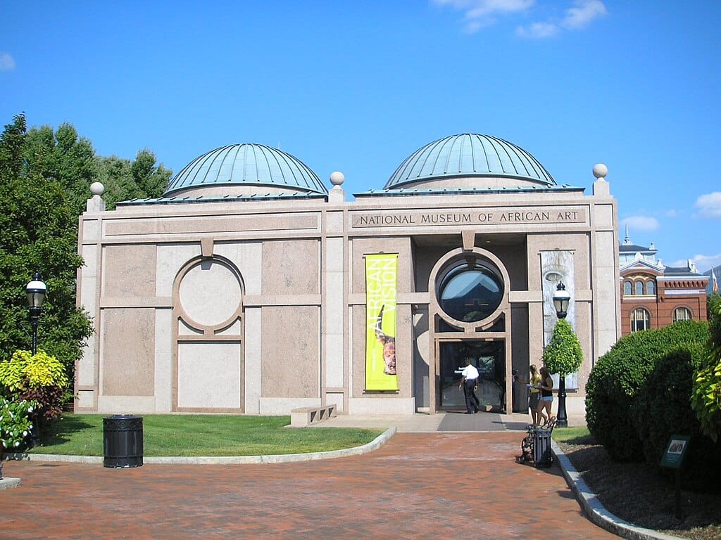 Exterior of a brown-ish building with two blue domes at the top, surrounded by greenery.