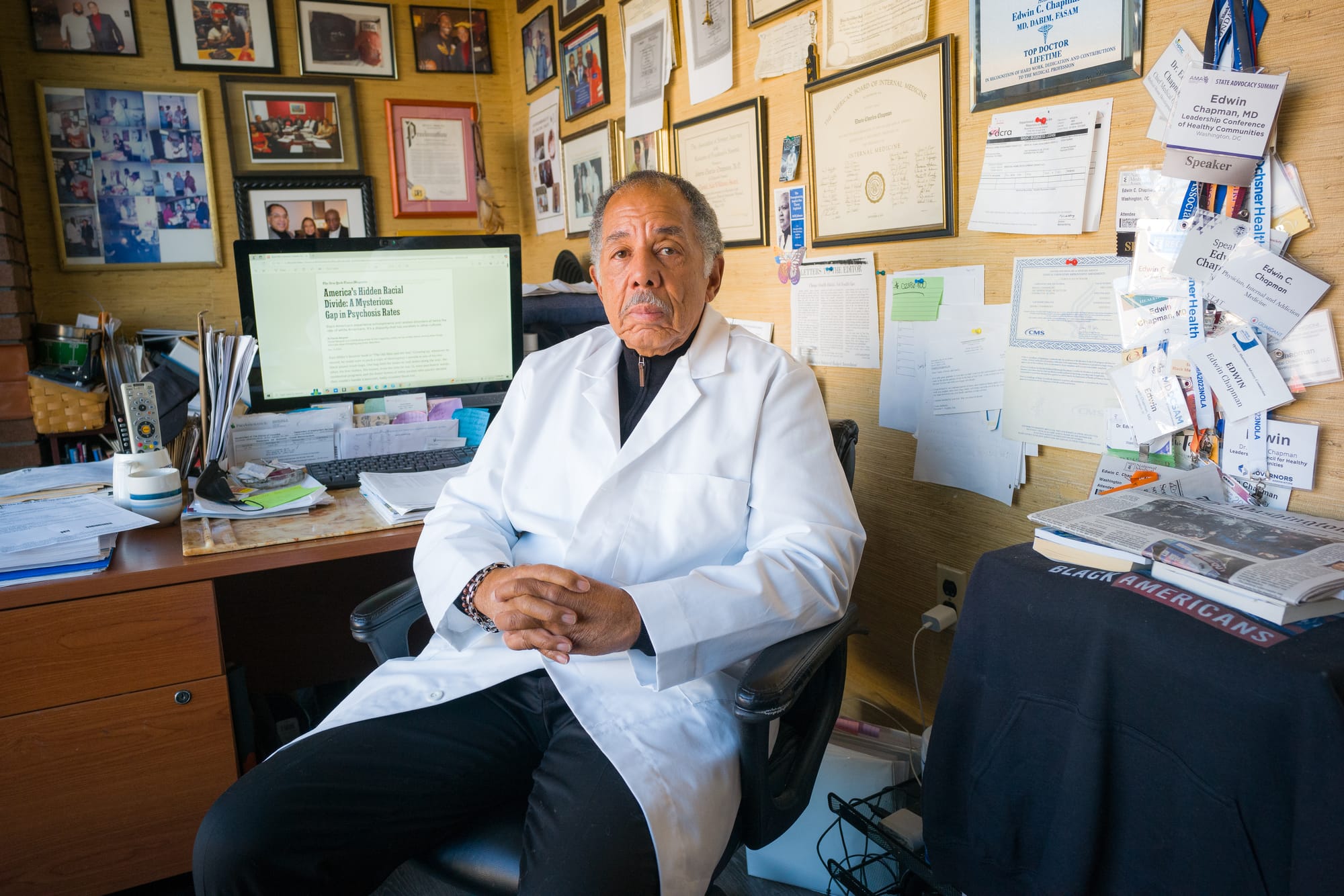 A doctor in a white coat sits in his office, which is covered wall to wall in framed awards and pictures. 