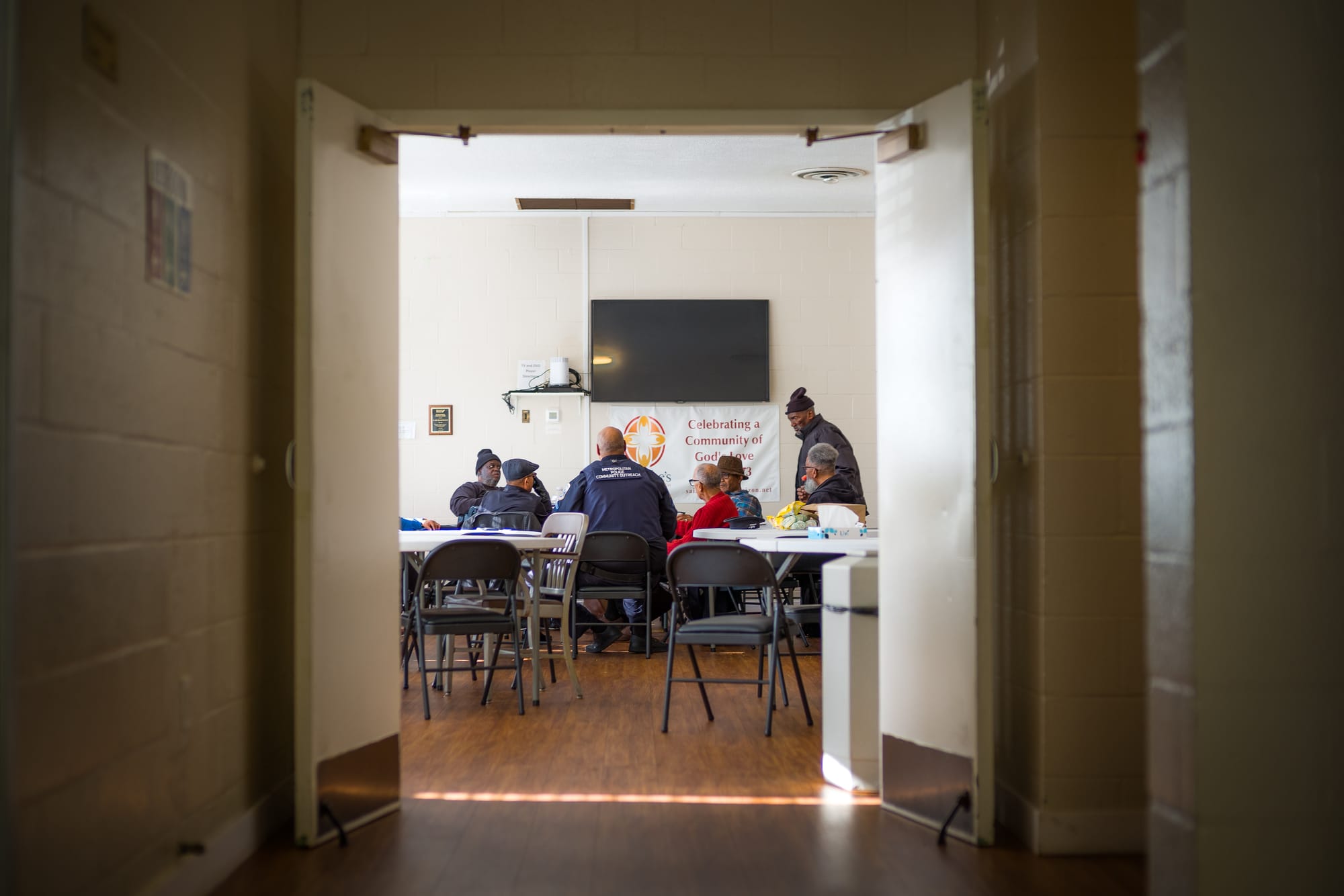 Two doors open from a hallway into a room full of chairs and tables with seven men talking.