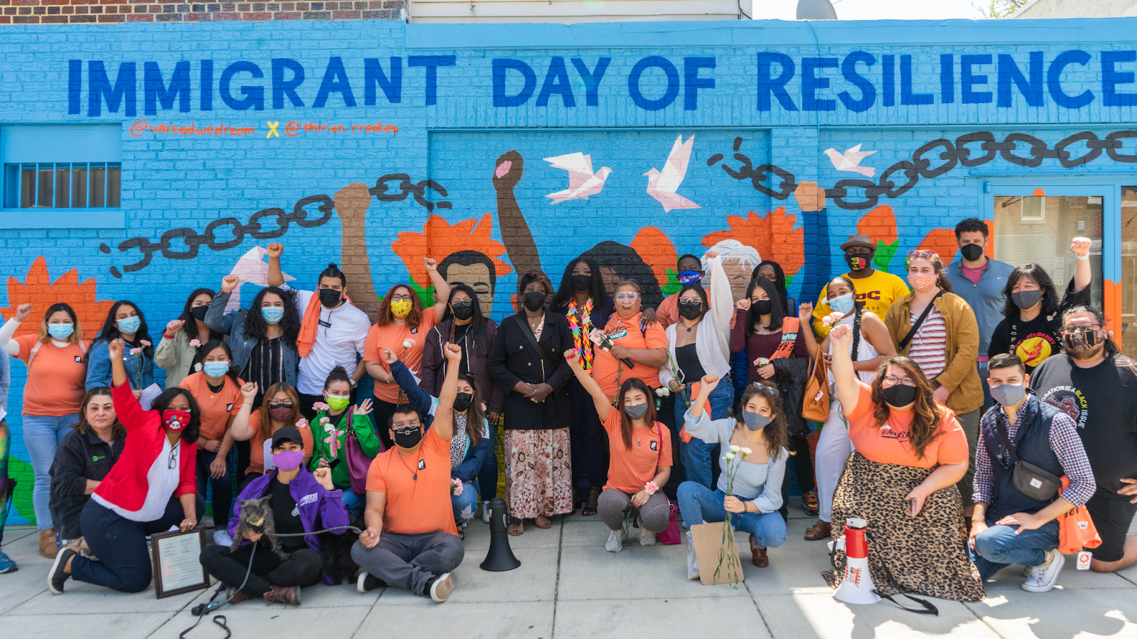 Ward 4 Councilmember Janeese Lewis-George holds a physical copy of the D.C. Council ceremonial resolution declaring April 15 as Immigrant Day of Resilience. She crouches in front of a mural that says the same, raising her fist alongside immigrant youth gathered there.