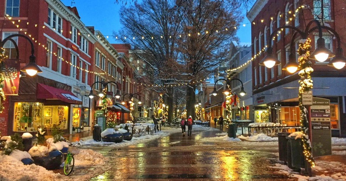 A snowy street at dusk, with lights wrapped around lamp posts and across a brick road. 