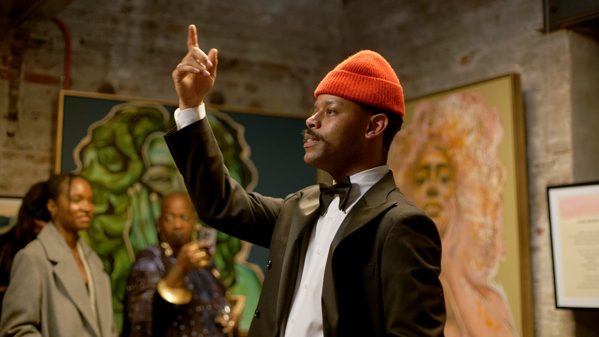 A man in a suit, bowtie, and orange at points his finger to the ceiling, appears to be speaking.