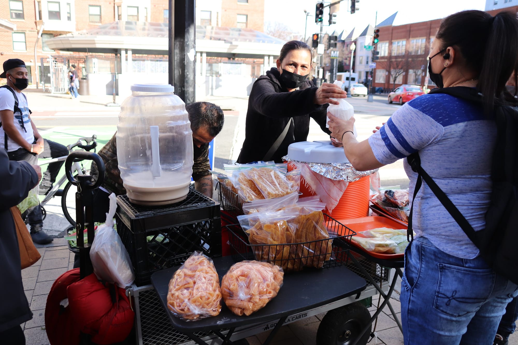 A vendor hands a package of food to a customer on 14th Street. Her cart is visible and full of snacks and a nearly empty jug of atole.