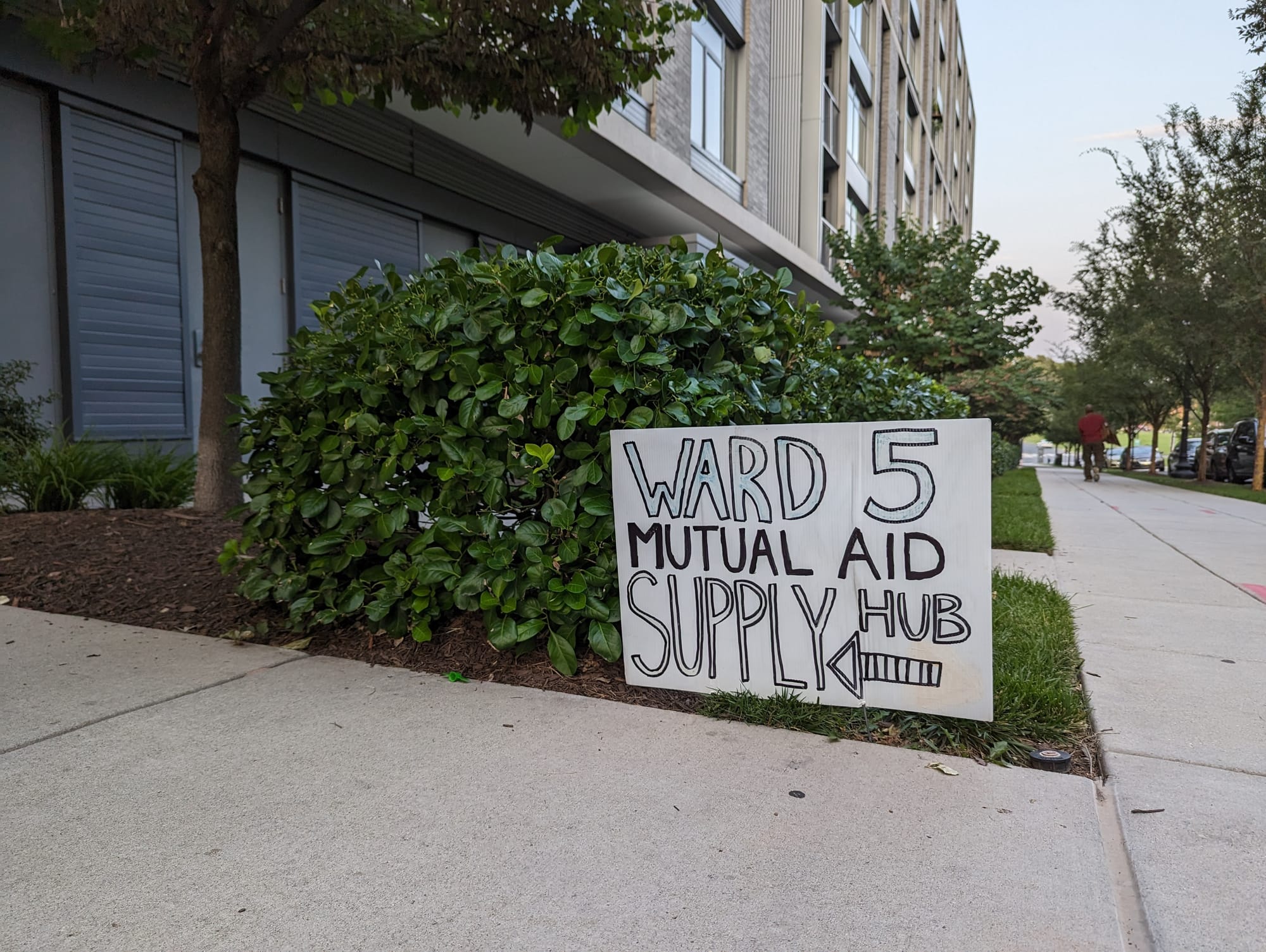 A white sign that reads “Ward 5 Mutual Aid Supply Hub” rests against bush next to a sidewalk.