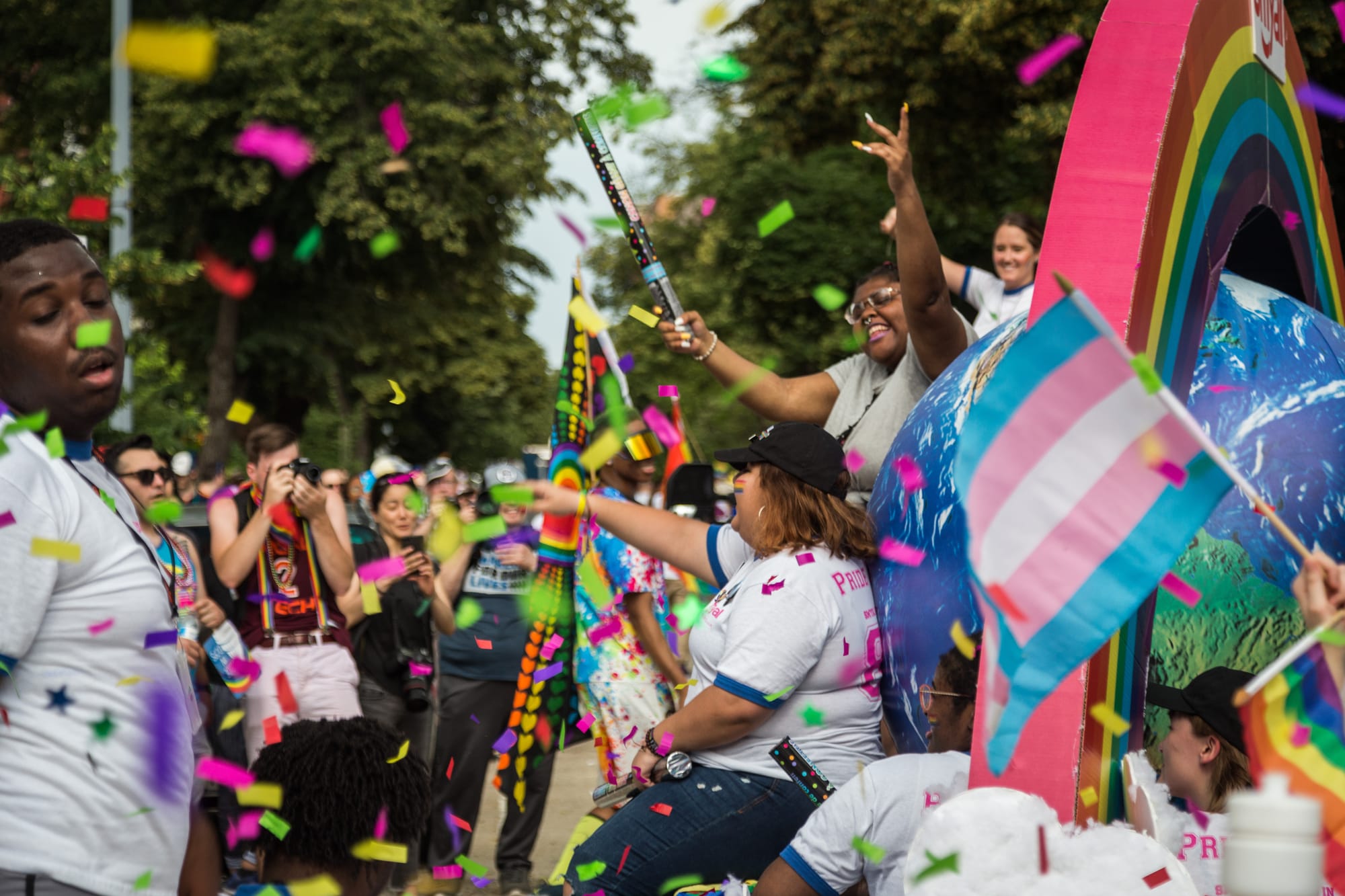 A colorful crowd celebrates Pride in rainbow confetti, with people on floats and dancing. A trans pride flag sits to the right of the frame.