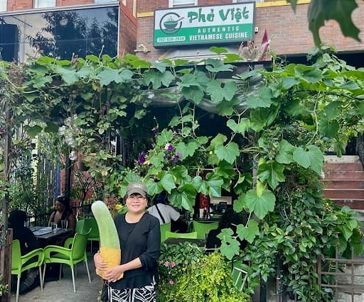 A woman stands in front of a restaurant with dense foliage covering the patio, holding a large vegetable in her arms.