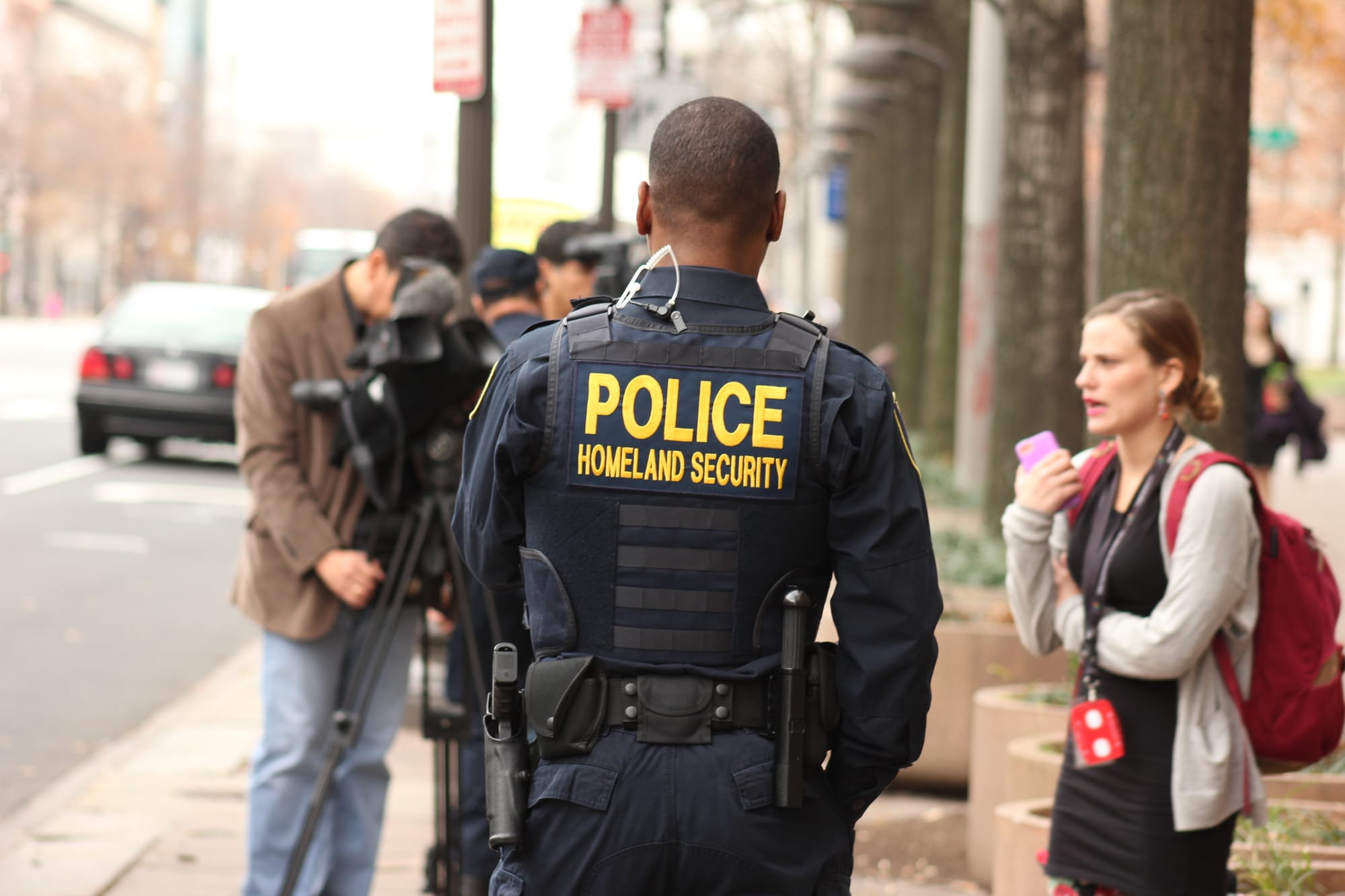 A photograph of a police officer on a sidewalk with a woman on her phone and a camera crew in the background.