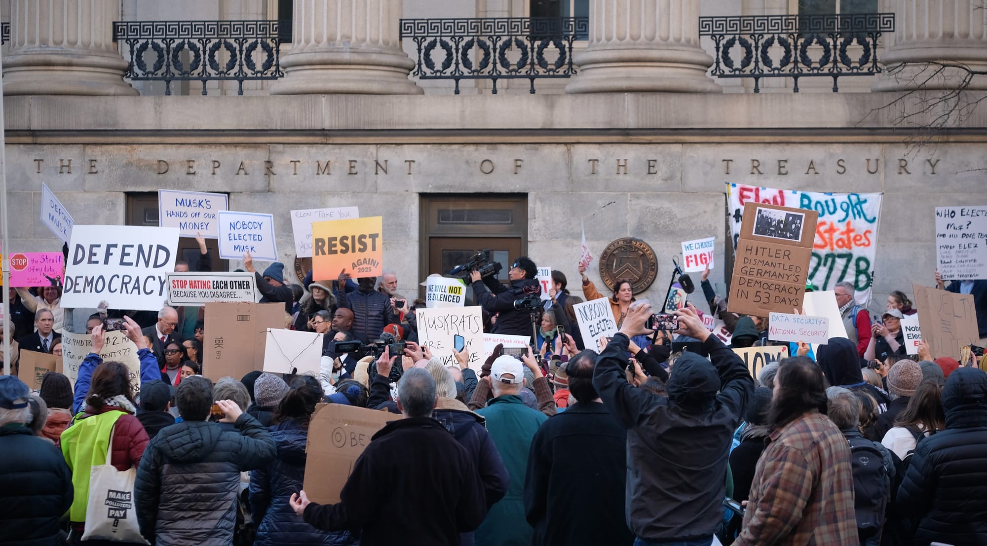 A large crowd of protestors outside the Treasury hold signs with messages including: "Defend Democracy" and "Resist."