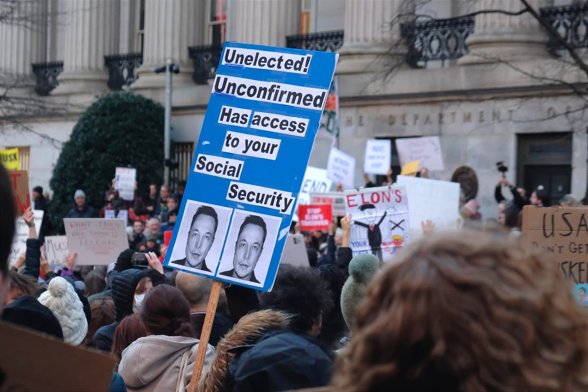 A protestor holds a sign reading: "Unelected, unconfirmed, has access to your social security," with two pictures of Elon Musk.