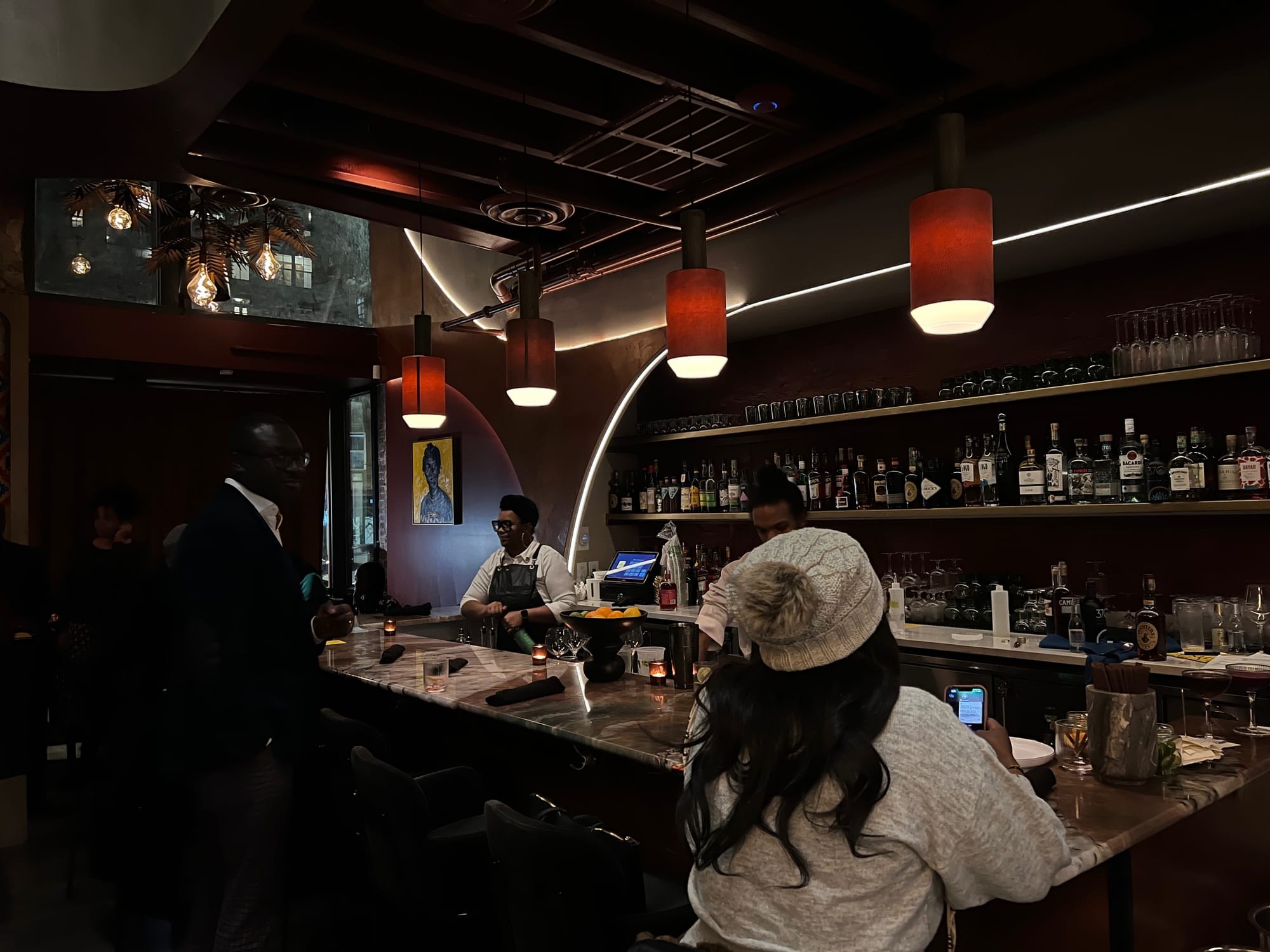 A dimly lit bar. A woman sits at the counter, while bartenders fix drinks. 