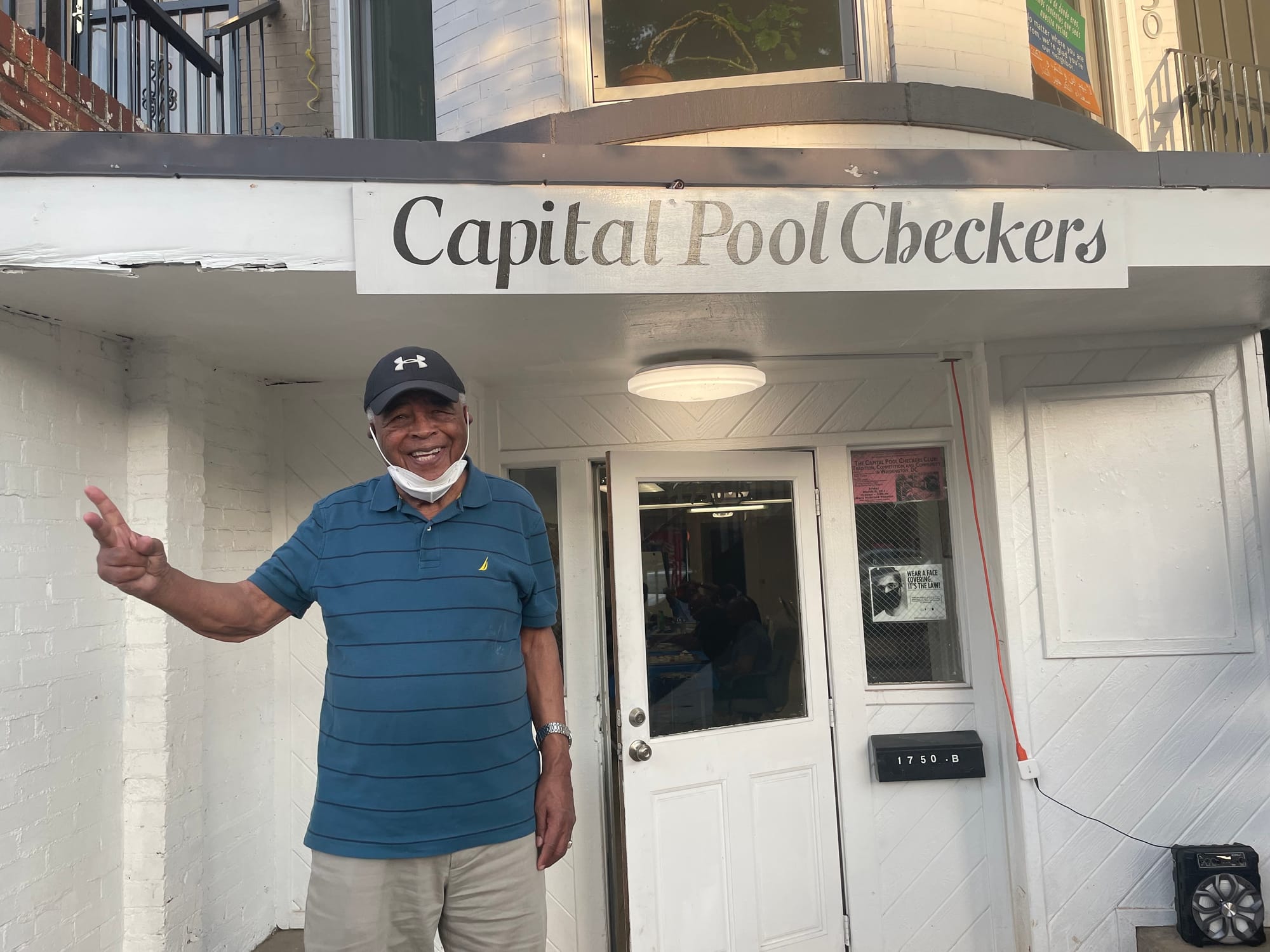 An older Black man holds up a peace sign in front of a building that reads "Capital Pool Checkers." 