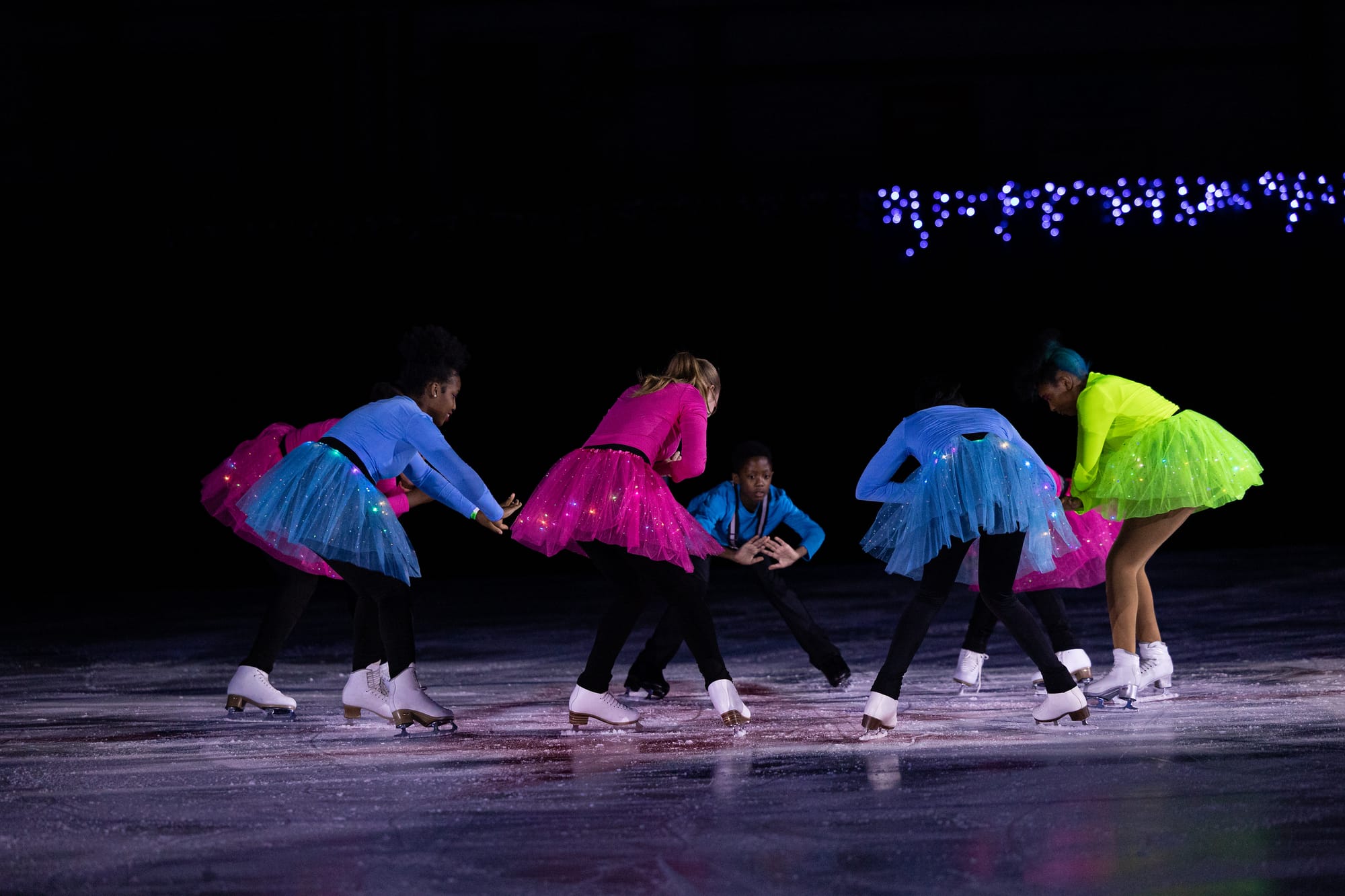 Young ice skaters perform on an ice rink in a circle with colorful, sparkly, neon outfits.