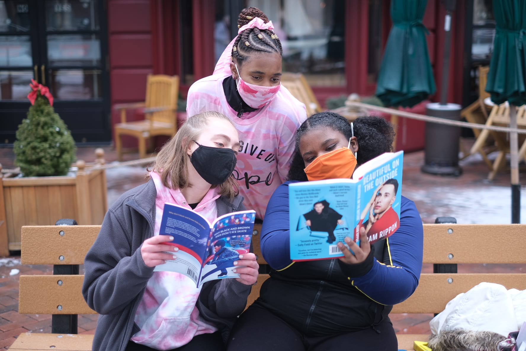 Three young ice skaters read books together on a bench outside.