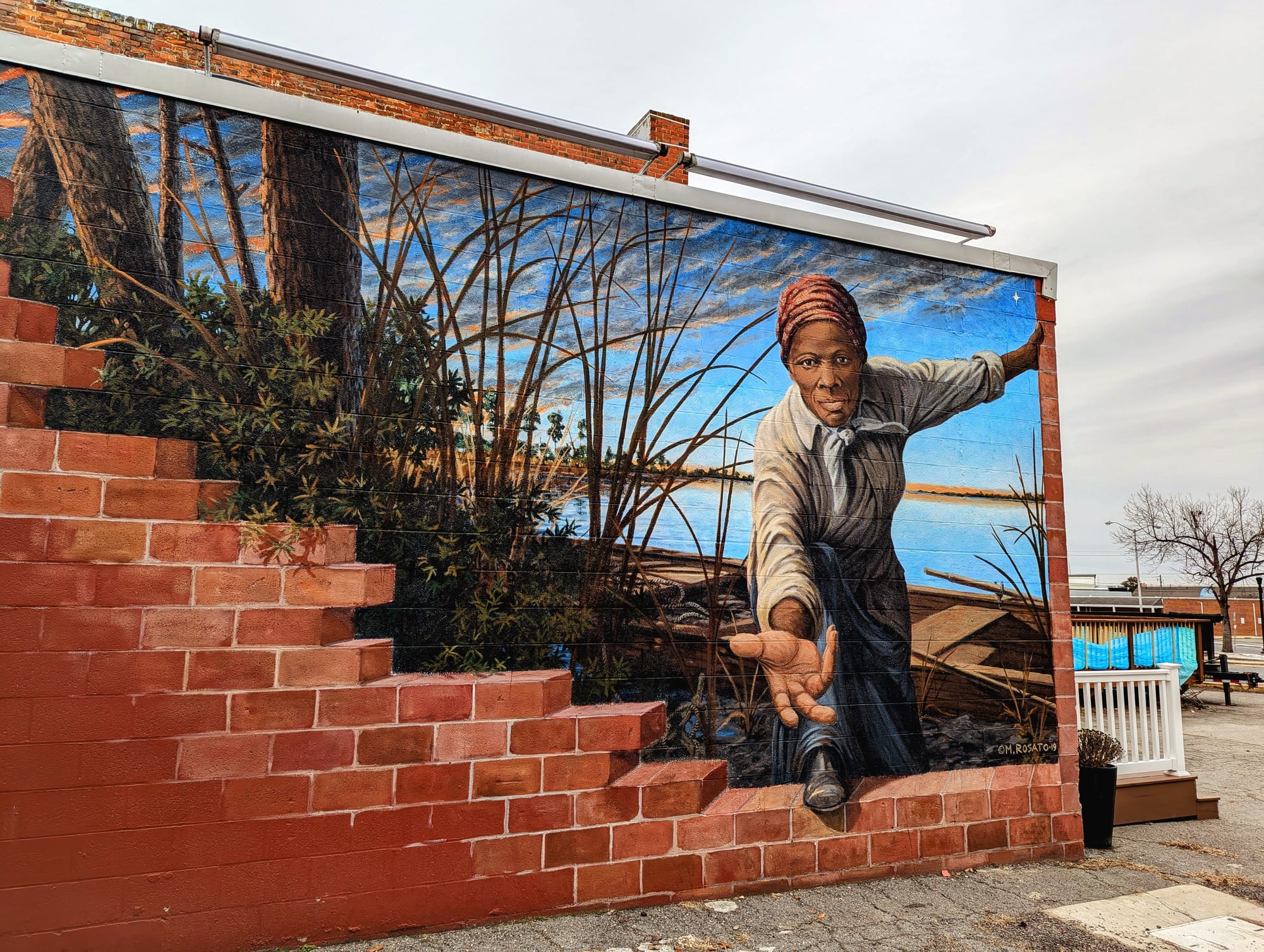 Photo showing a mural by Michael Rosato (https://michaelrosato.com) depicting Harriet Tubman, with the Easter Shore landscape behind her, stepping over a brick wall to offer a hand to the viewer.