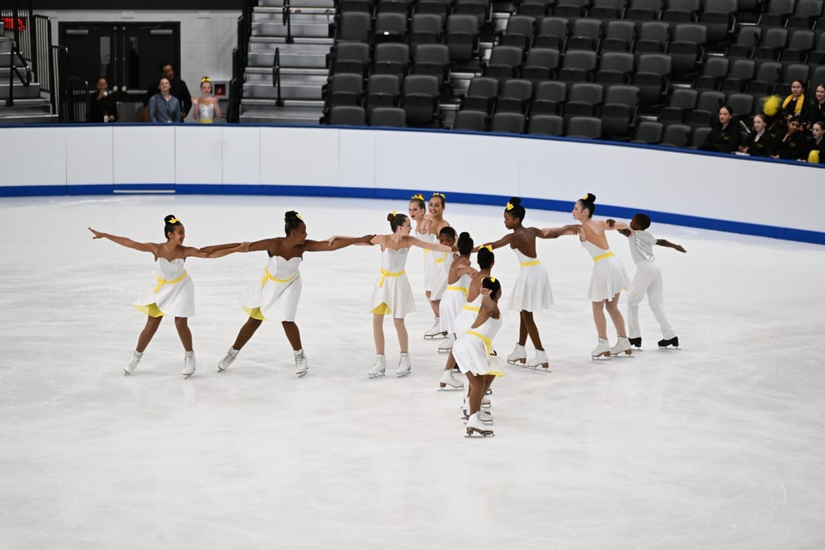 An image of a line of figure skaters linking arms, all in white uniforms with yellow accents.