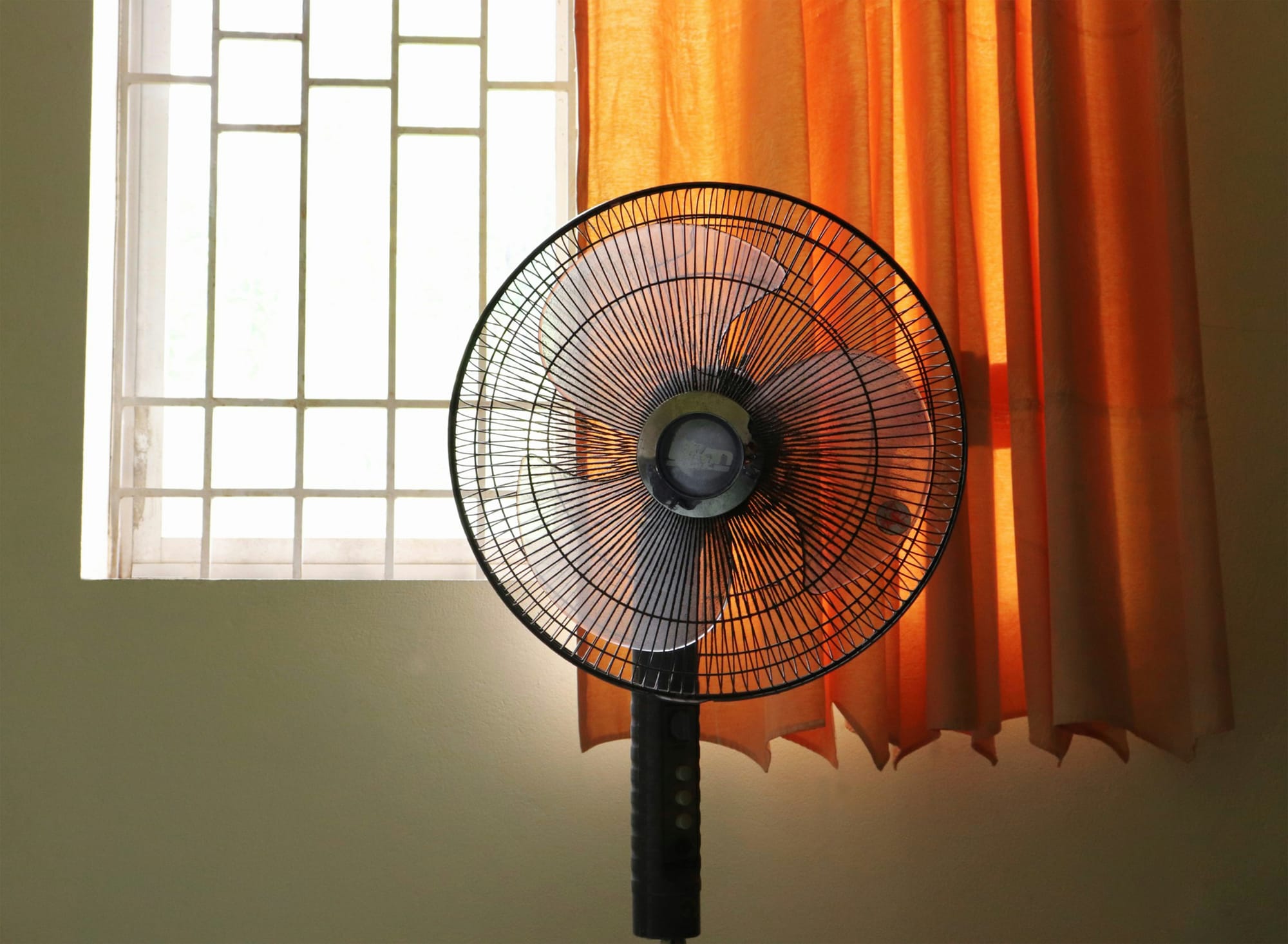 A standing fan in front of a window with an orange curtain