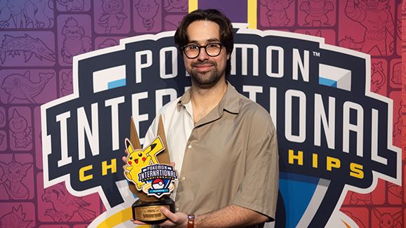 A man holds a Pokémon trophy in front of a background image for the International Championships
