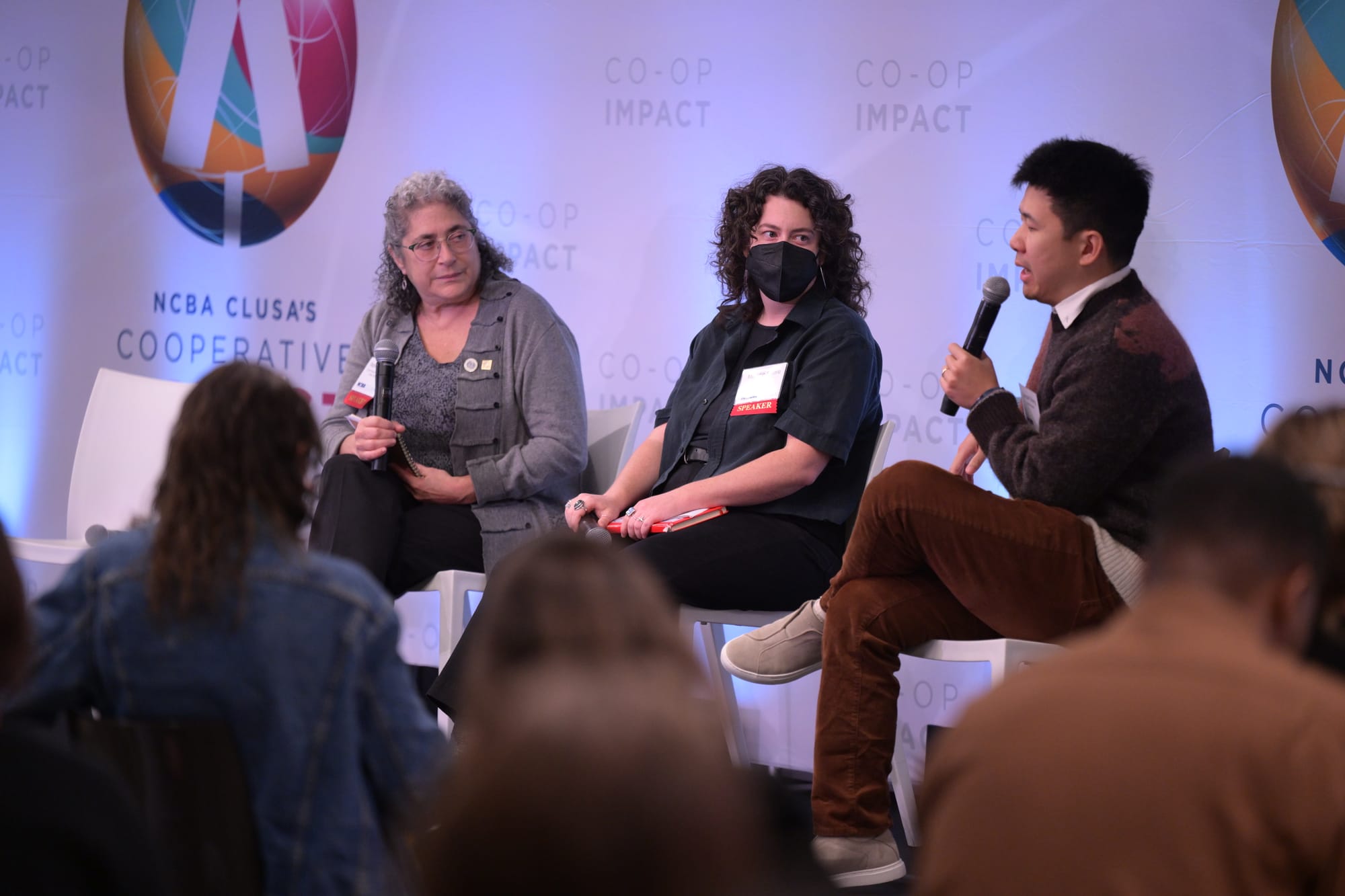 Three people sit in chairs on stage, with one speaking into a microphone.