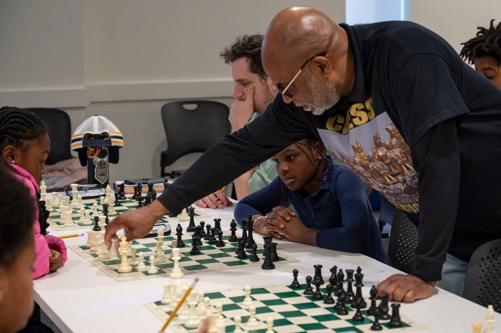 A photo of a man leaning over three chess boards, with several students watching.