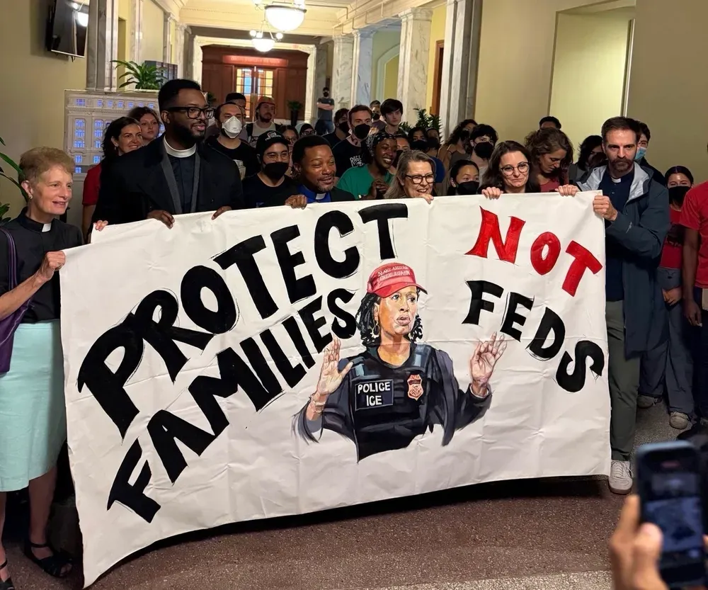 A group of advocates stands in front of a poster depicting Mayor Bowser in a police vest that reads "ICE." The mayor has a red hat on. The poster reads, "protect families, not feds."