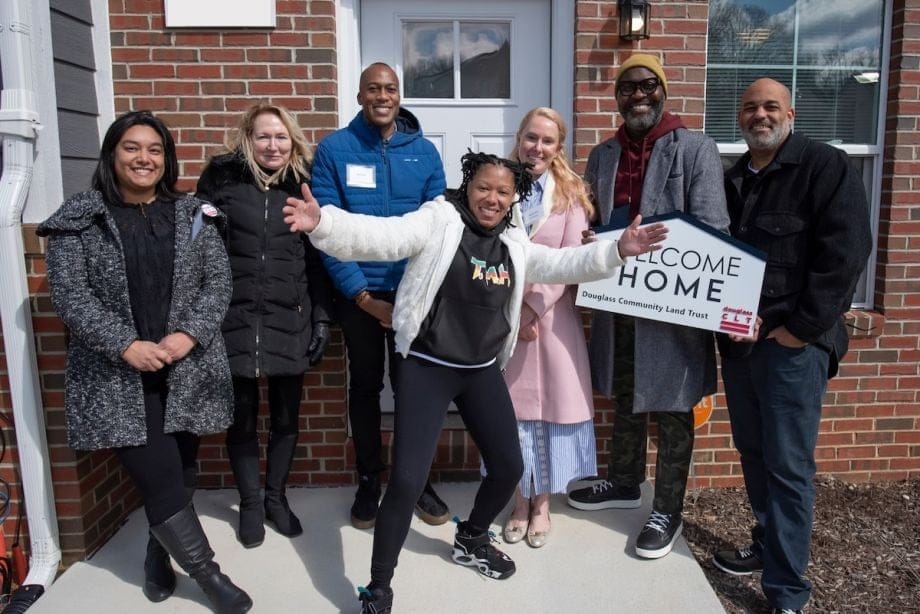 A group of people of different races stand in front of a brick house, holding a sign that says WELCOME HOME.