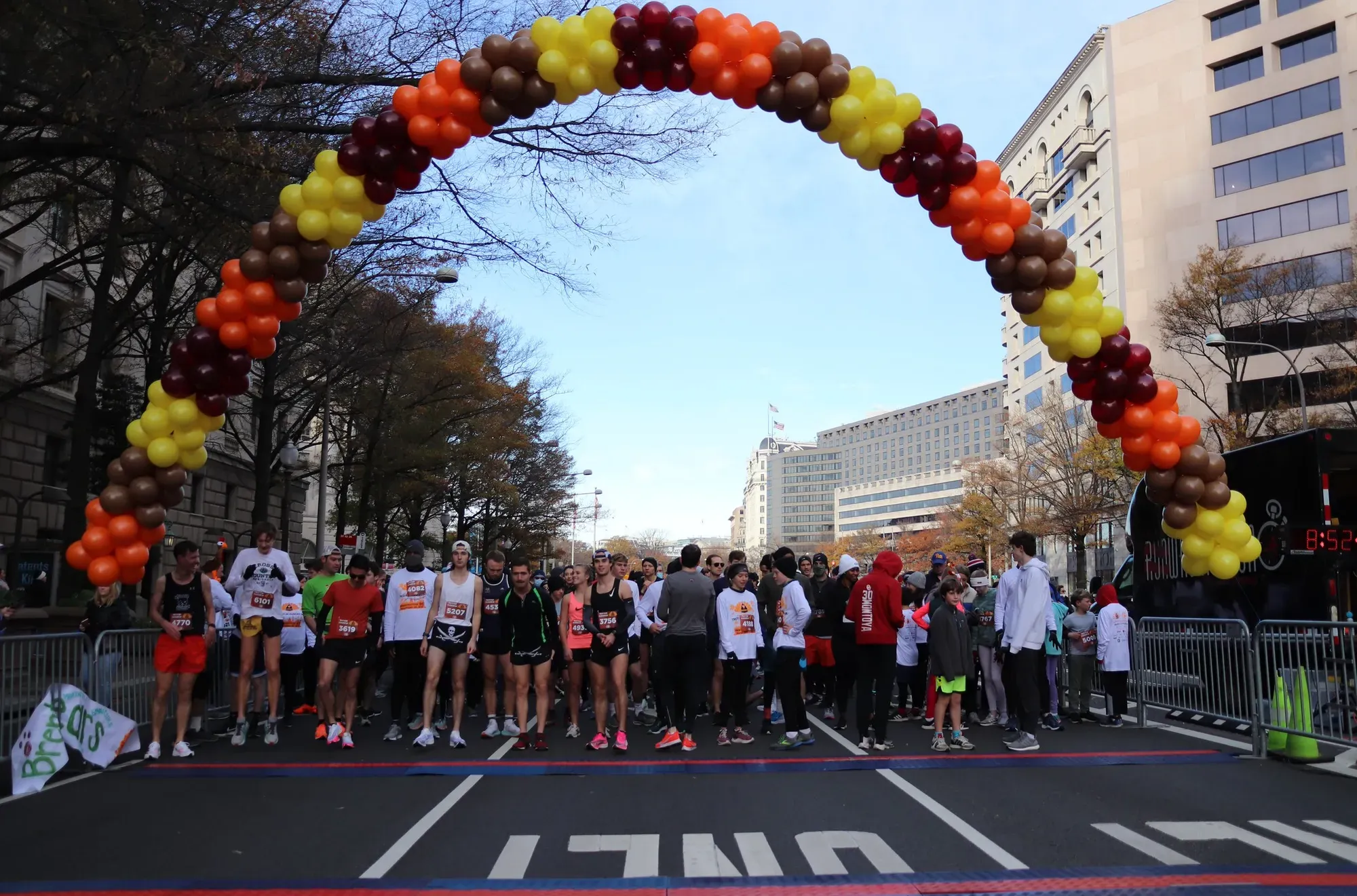A crowd of runners lined up at a starting line, under a Thanksgiving-themed arch of balloons.