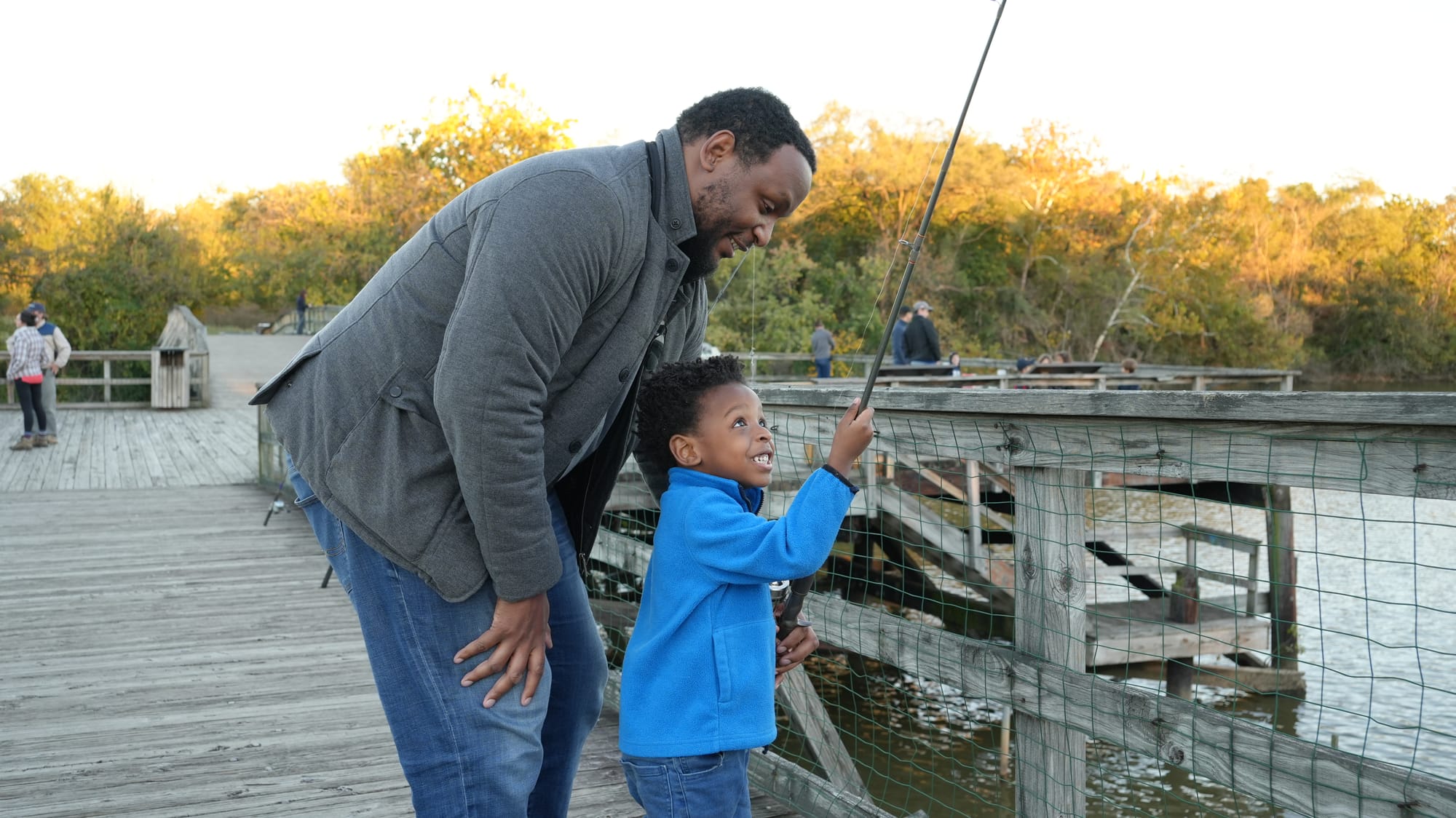 A Black man helps a young Black boy who is holding a fishing rod on a dock in a river.