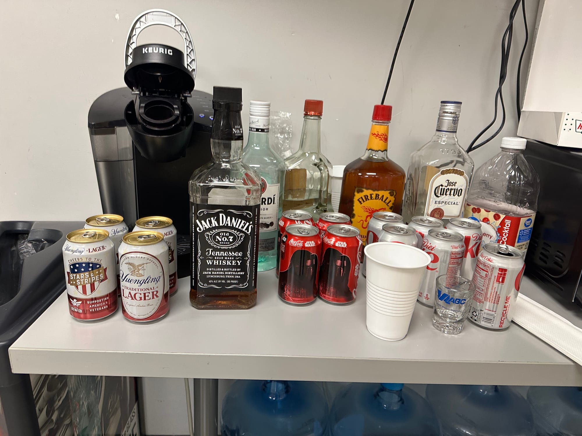 An office table with a Keurig and various types of alcohol on it: Beers, Jack Daniels, Bacardi, Fireball, and Jose Cuervo.