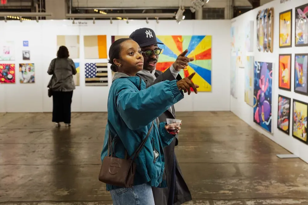 Two people in an art gallery point at a work that is out of frame to the right