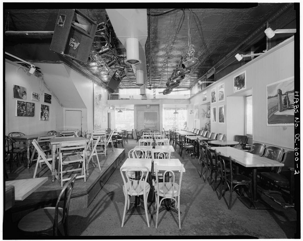 A black and white photo shows the empty inside of d.c. space — featuring tables and chairs arranged around a large room. 
