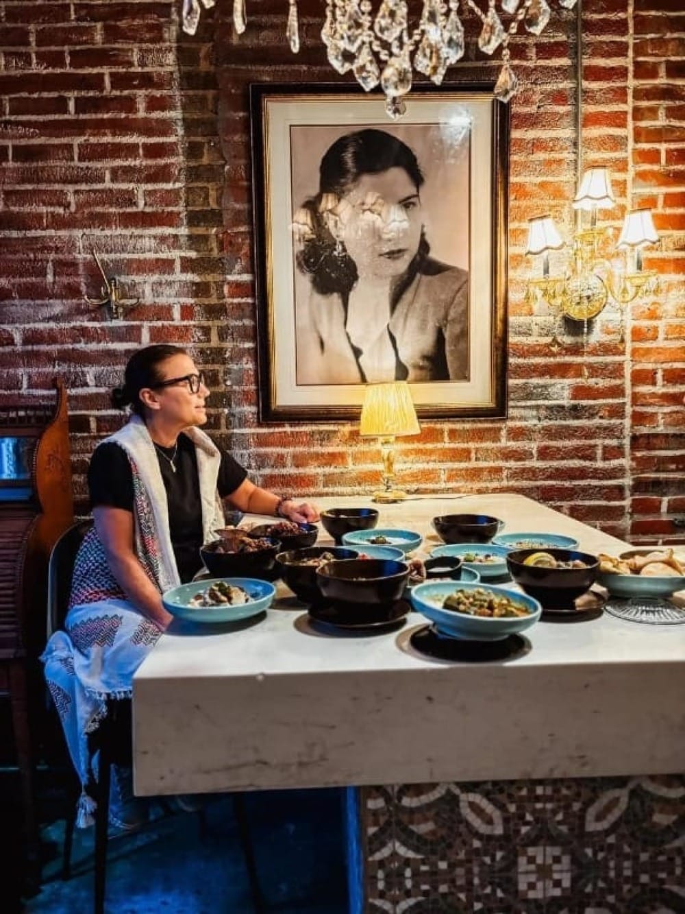 A woman seated at a table in front of a variety of dishes, under a black and white portrait. 