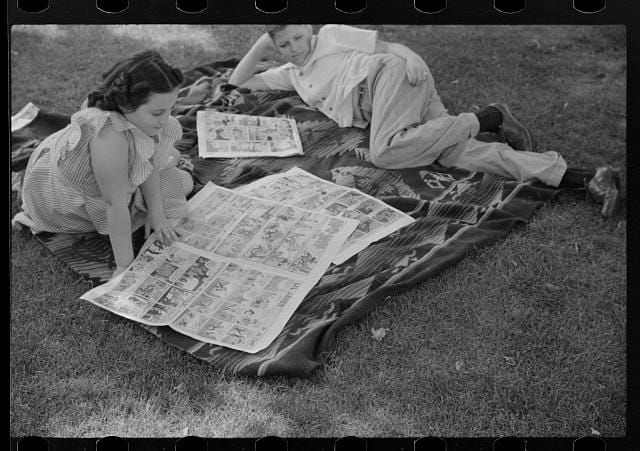Two children read comics on a blanket in a park.